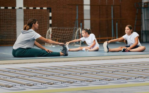 Sports teacher and students stretch during physical education class at school gym.