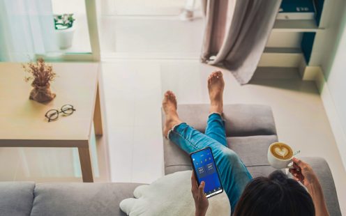 A woman sitting on a sofa and holding a smartphone with indoor temperature data displayed.