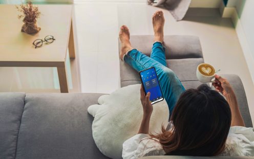 A woman sitting on a sofa and holding a smartphone with indoor temperature data displayed.