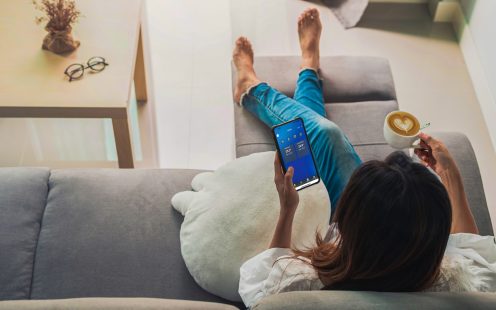 A woman sitting on a sofa and holding a smartphone with indoor temperature data displayed.