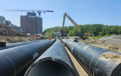 A close-up of three large-diameter stormwater pipes with a long-reach excavator working in the background.
